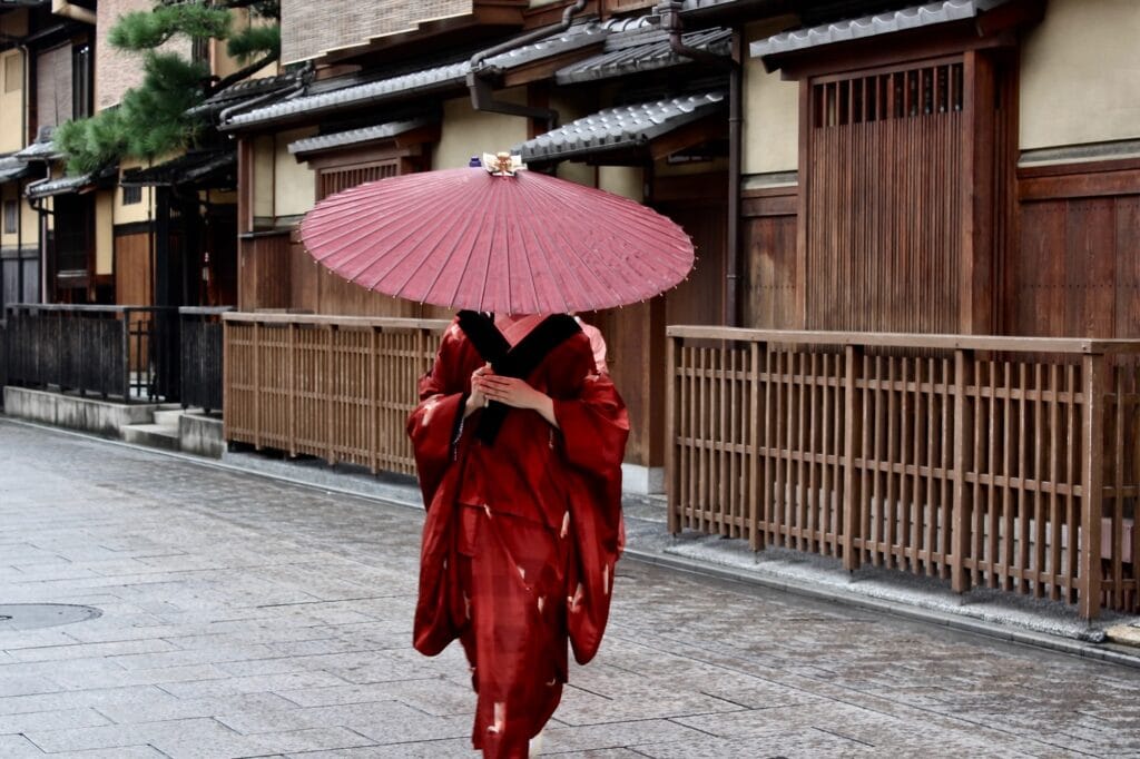 A geisha walks in kimono down a Kyoto street, a paper umbrella obscuring her face.