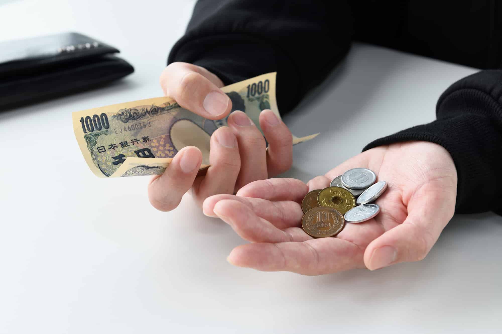A person holds onto Japanese yen bills and spare coins, their gestures appearing upset. An empty wallet lays on the table nearby.